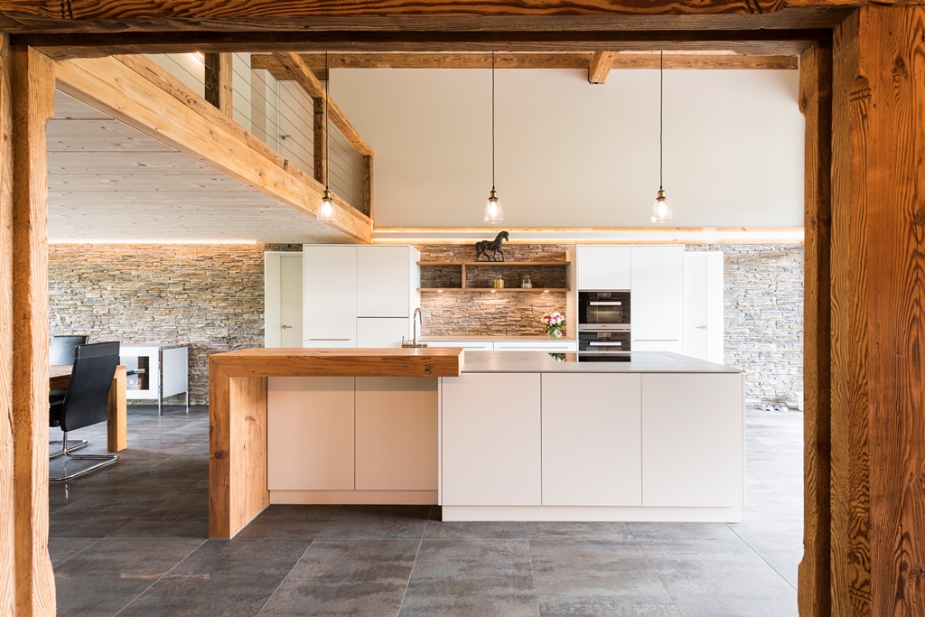 Der Raum wurde im Zuge eines Bauernhaus Umbaus gestaltet. Die moderne Küche in der Mitte des Raums verfügt über weisse Fronten und eine Kochinsel mit einer Stehbar aus Holz. Ein Boden aus grau-braunen Steinfliesen und eine Wand aus Stein verleihen dem Raum einen besonderen Charakter - geplant und realisiert durch die Kobi Architektur AG im Buechiberg (Solothurn)